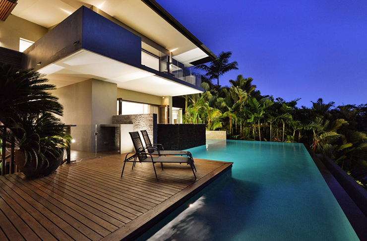 A three-story mansion lit up at night and surrounded by a wrap-around pool.