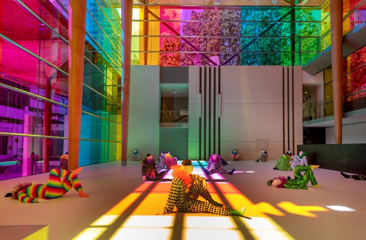 A whole lot of clown sculptures in varying poses sitting on the floor, almost bathing in rainbow light. 