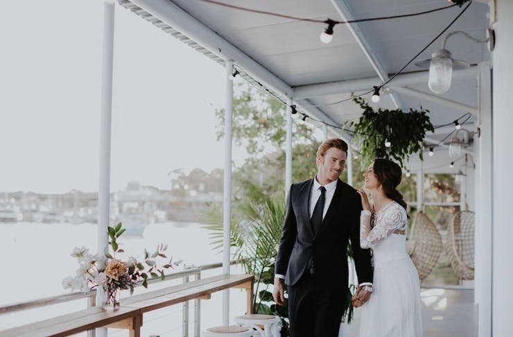 A bride and groom embrace on a waterfront balcony, under festoon lights. 