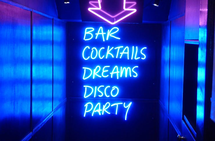 A purple neon arrow pointing down some stairs with the words bar, cocktails, dreams, disco and party. 