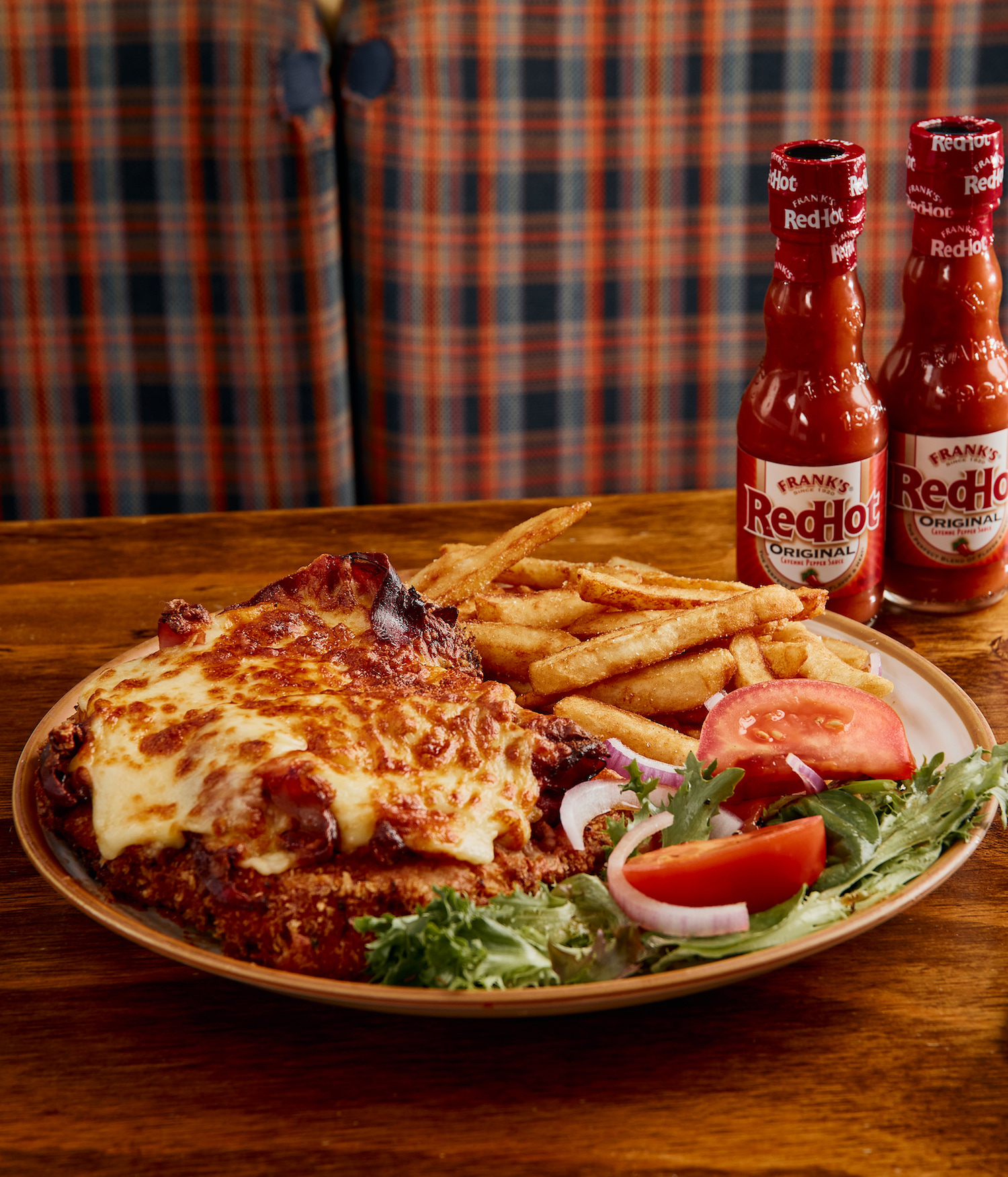 A cheesy chicken parma sits next to two bottles of Franks RedHot Sauce