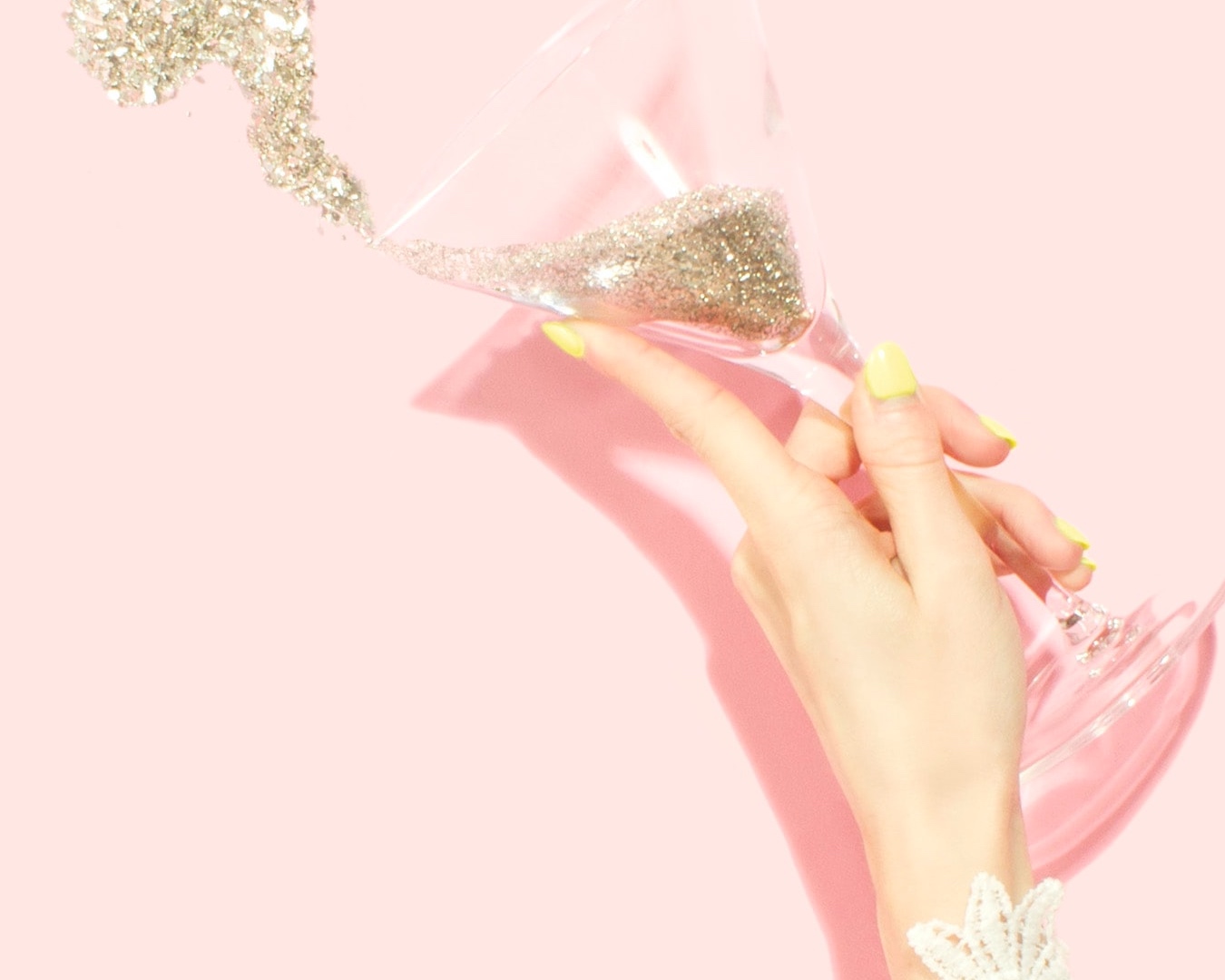A hand holding a champagne glass filled with glitter that’s splashing out onto a baby pink background. 