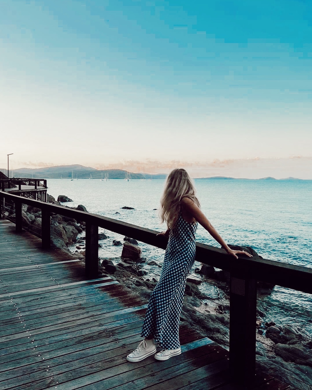 A woman standing on a boardwalk wears a gingham dress and white converse while looking out to sea. 