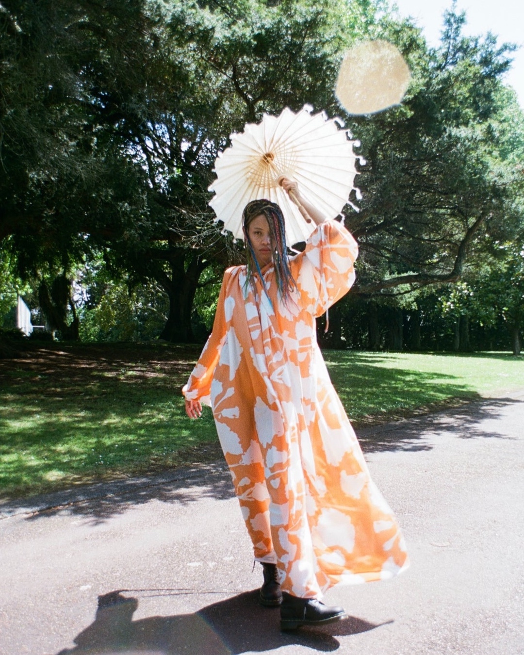 A woman with multicolored braids, wearing a flowing orange and white dress, holds a white parasol in a park. 