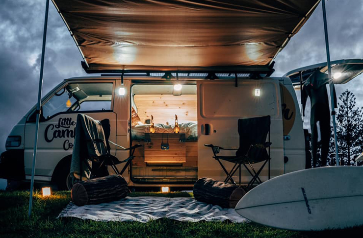 A shot at dusk of a lit up camper van with camping chairs positioned under an open awning.