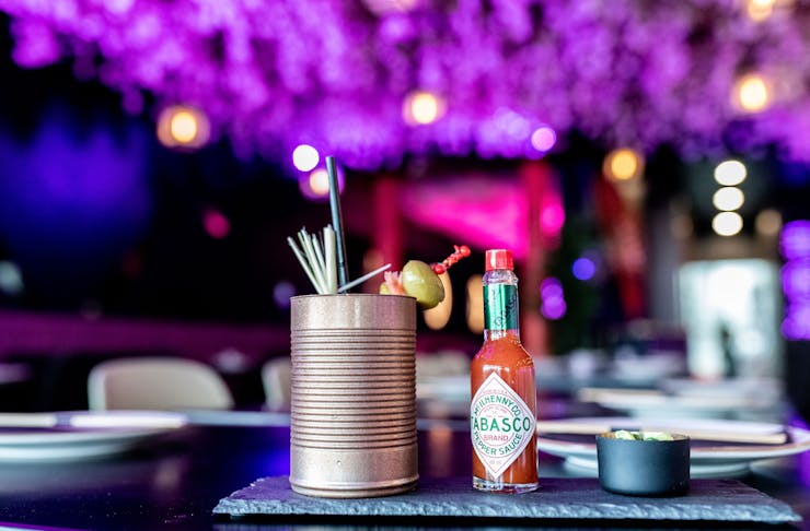 A Bloody Mary style cocktail served in a tin can with a bottle of Tabasco beside it, against a neon-purple backdrop.