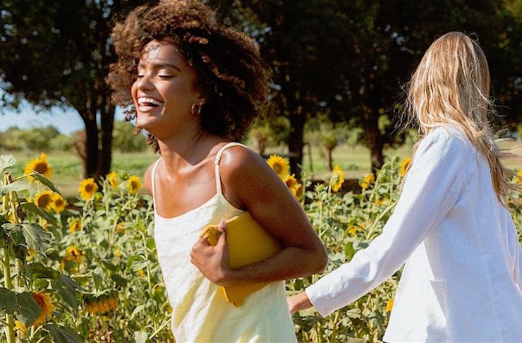 Two woman run in a field of sunflowers smiling and laughing.
