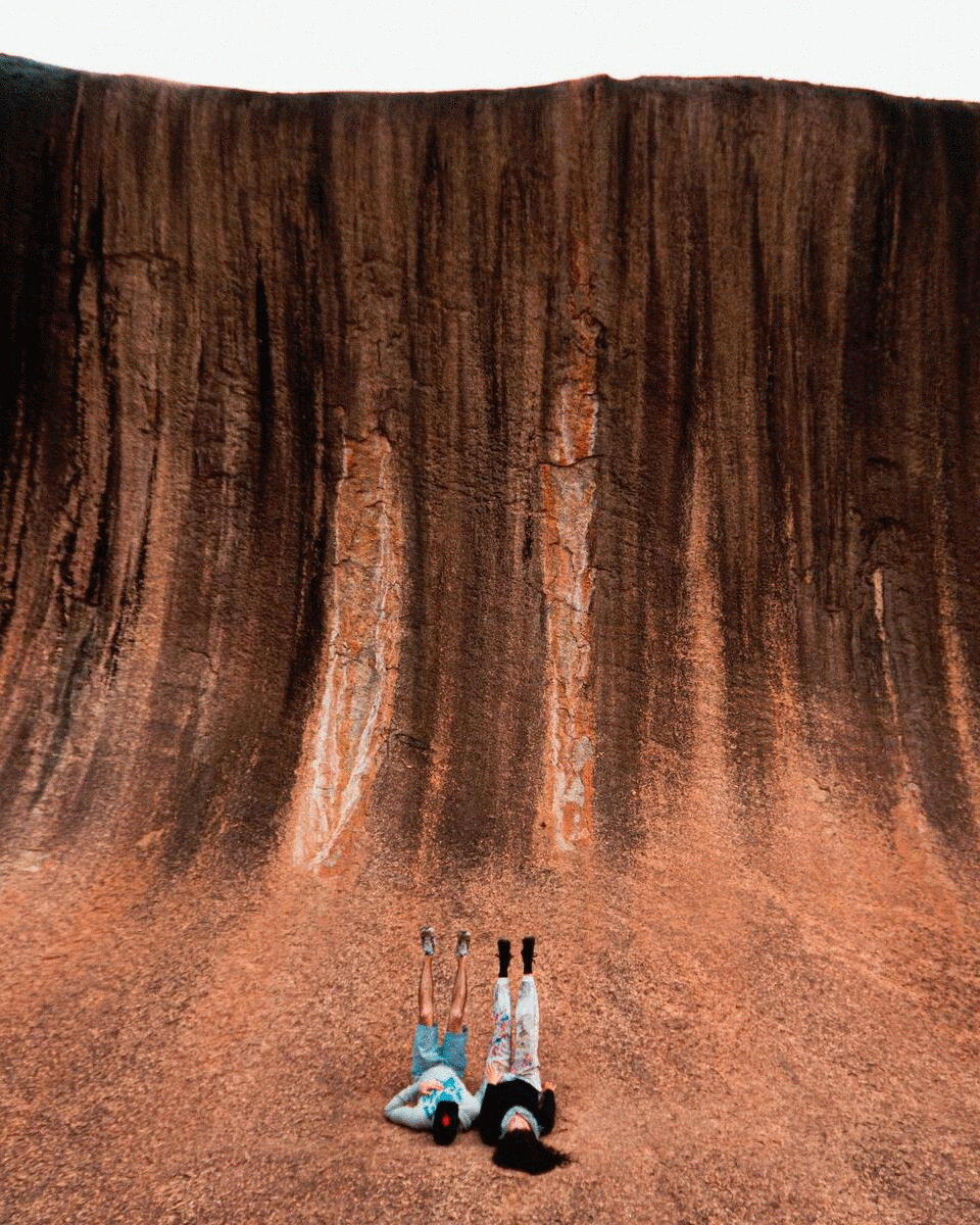 Two people lie no their backs with their legs in the air at the wave rock in WA. 