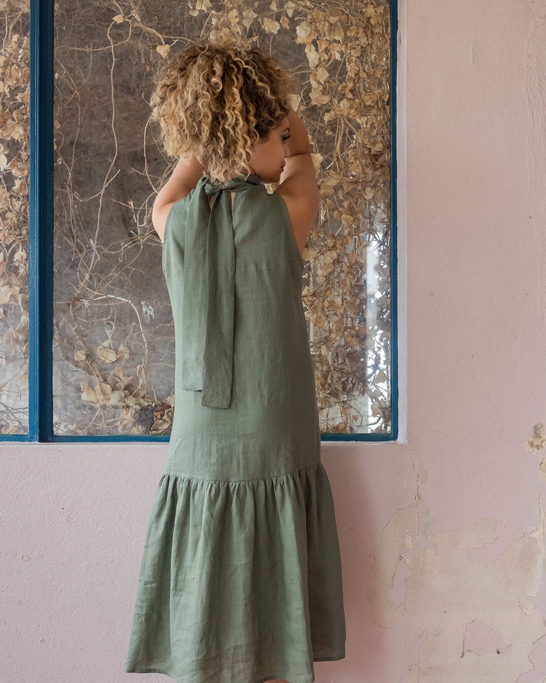 We see a woman from behind, she holds her hair up and looks aside while wearing a green linen dress. 