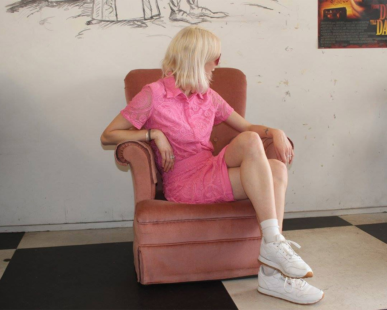 A woman sits in a dark pink armchair wearing a light pink lace dress as she looks off camera. 