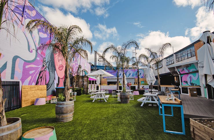 An outdoor garden bar with palm trees, lawn, and pink coloured art murals.