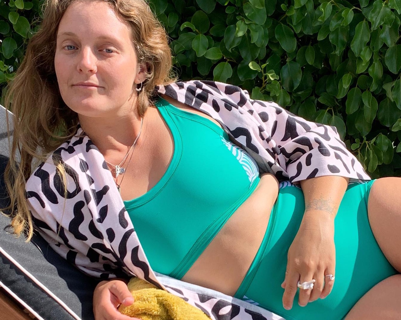 A woman lounges on a chair in a green bikini and a cover up as she looks at the camera. 