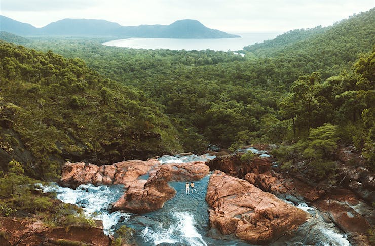 Two people wade at the edge of a waterfall overlooking lush rainforest.