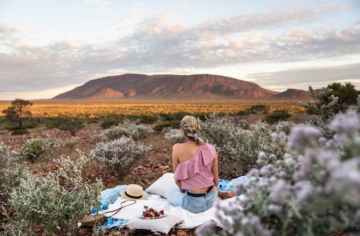 A woman sits on a picnic blanket as she gazes out towards the Western Australian outback.