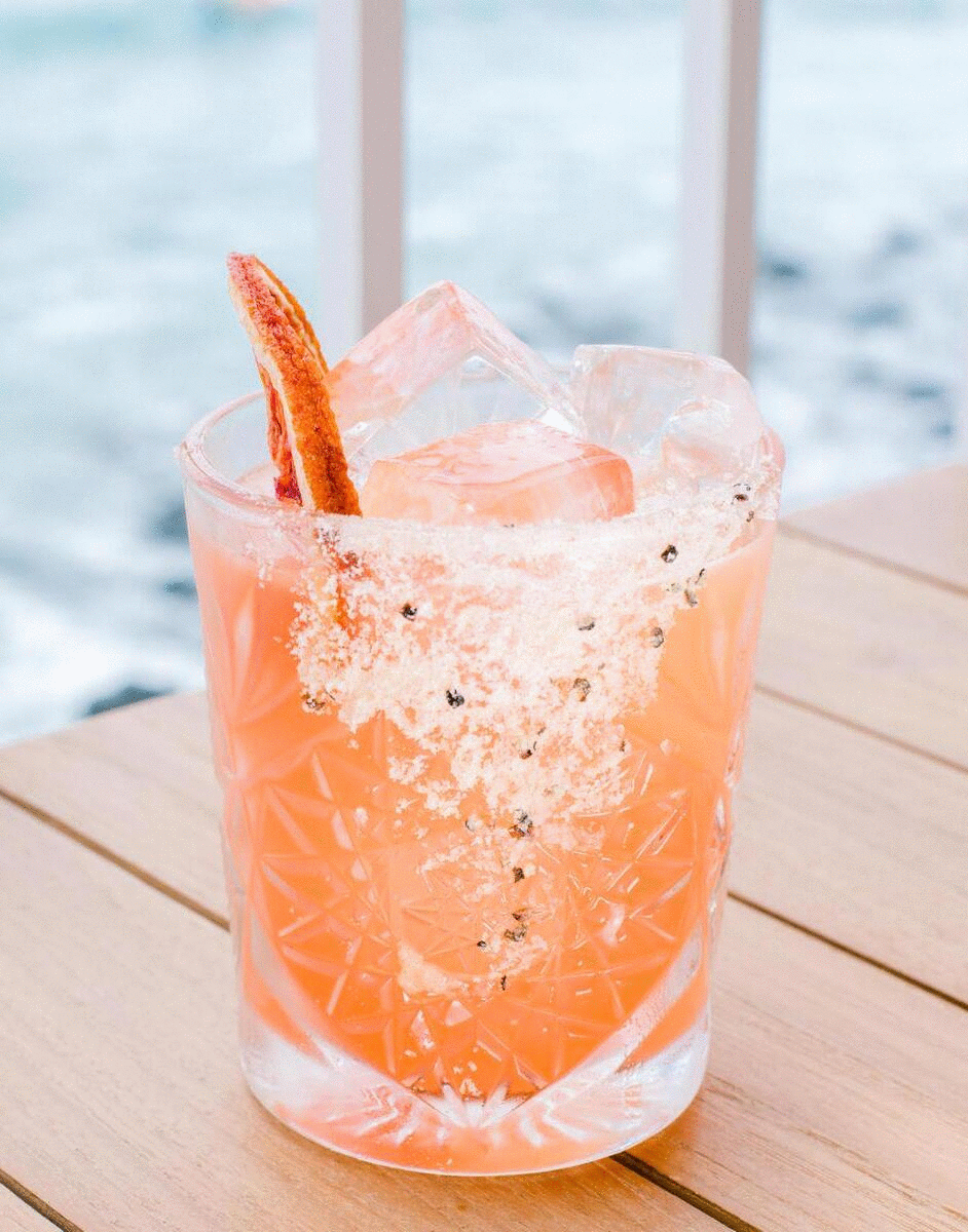 A zesty orange cocktail sits on a wooden bench near the ocean.