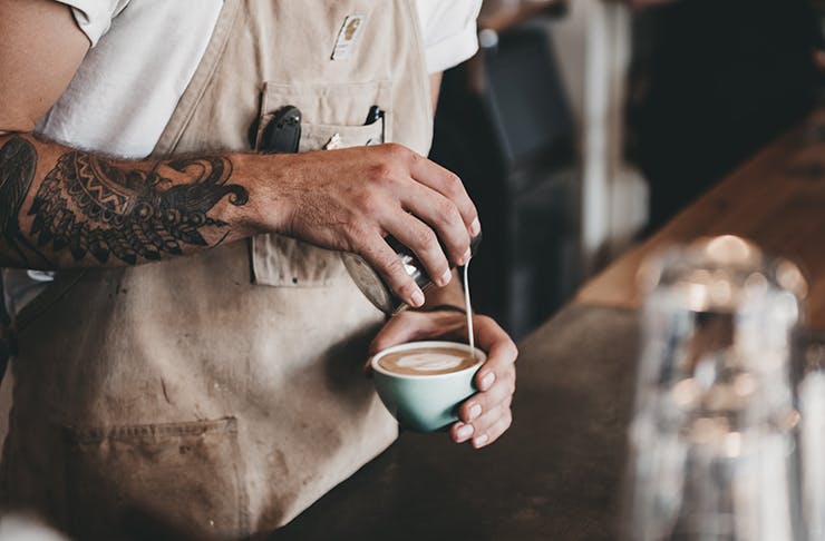 A tattooed male barista holding a coffee while pouring milk into the cup.