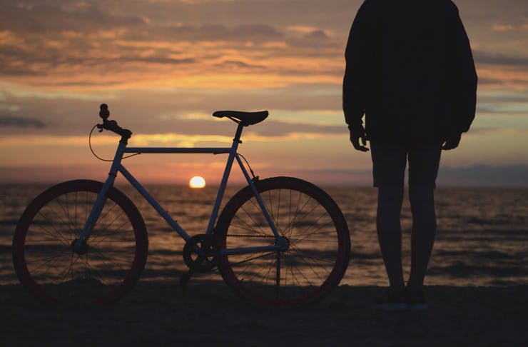 A man standing beside his bike at the seaside at sunrise. 