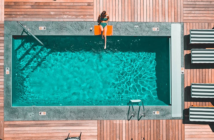 An aerial view of a pool surrounded by wooden decking. A woman is sunbathing on the deck.