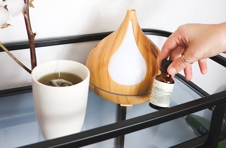 A person holding an essential oil next to a diffuser and a cup of tea.