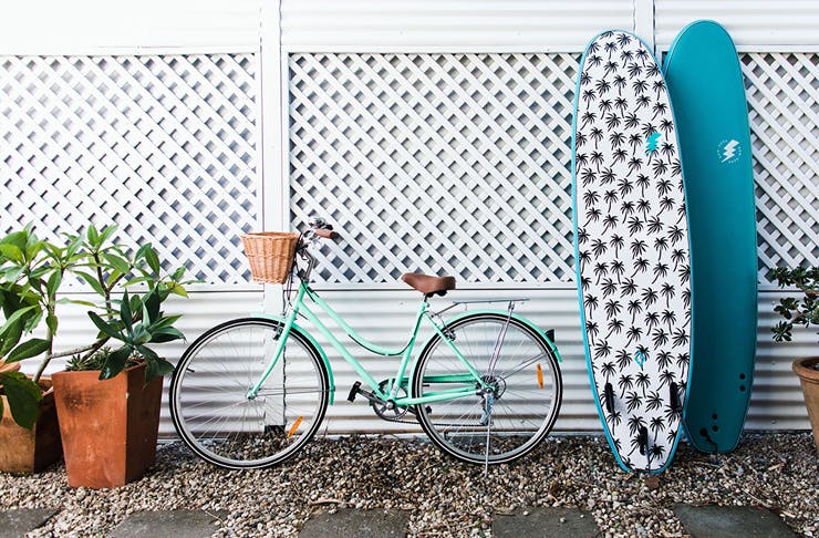 Two surfboards and a turquoise cruise bike lean up against a white wall.