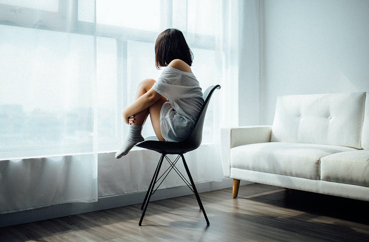A girl looks out of a window as she sits curled up on a chair.