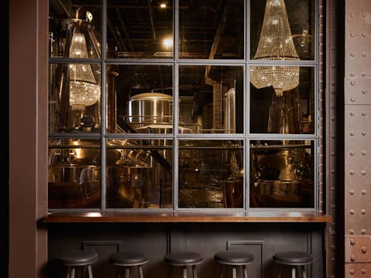 Wooden bar stools beneath a window looking into a gin distillery with copper pot stills. 