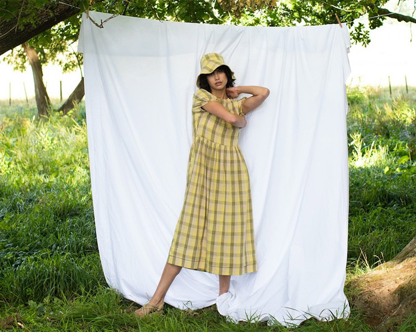 A woman in a yellow gingham dress and bucket hat stands in front of a sheet in a field. 