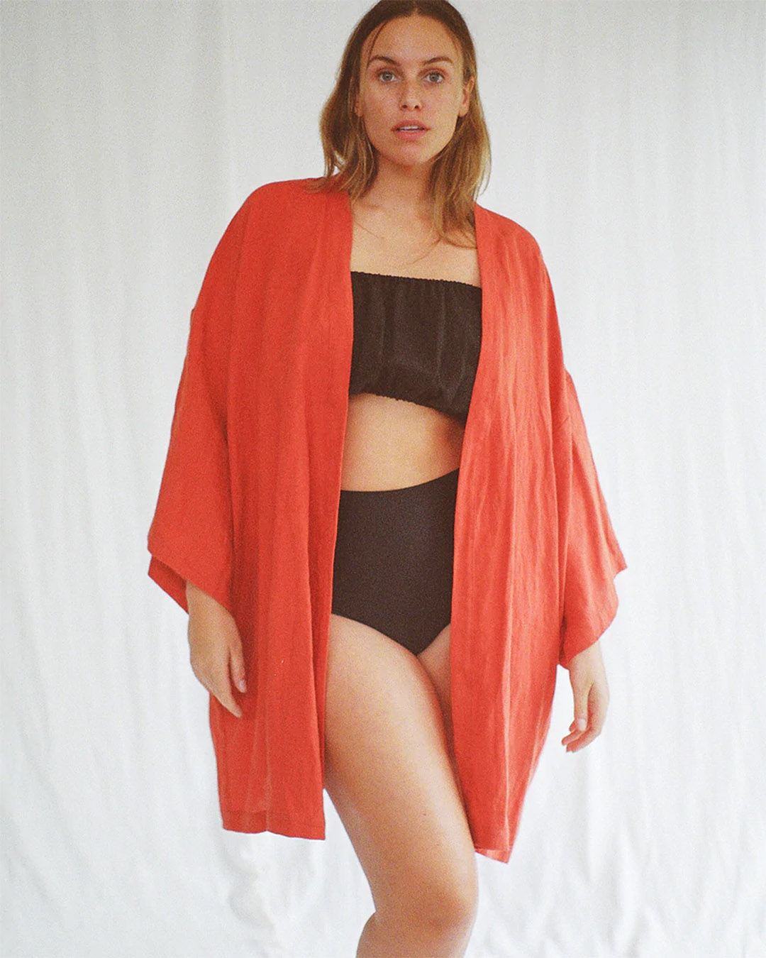 A woman wearing an orange kimono style dress over brown high waisted separates. 