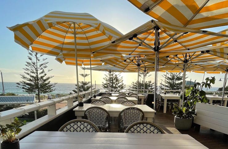 white tables topped with yellow beach umbrellas on a deck looking out to the ocean as the sun sets 
