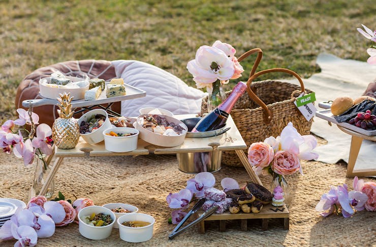 A display of food and beverages from a picnic hamper on a blanket over the grass