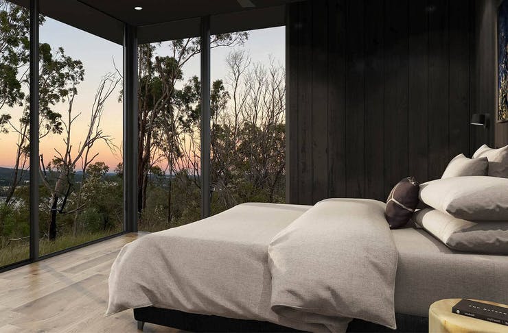Queen bed in front of large windows overlooking bushland and a sunset.