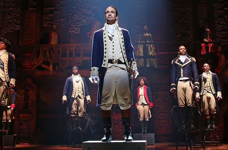 The character of Alexander Hamilton stands in the middle of the stage, with co-stars scattered around him.
