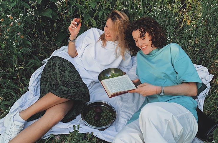 Two women laying on a picnic blanket in the grass laughing, holding a book and eating olives.