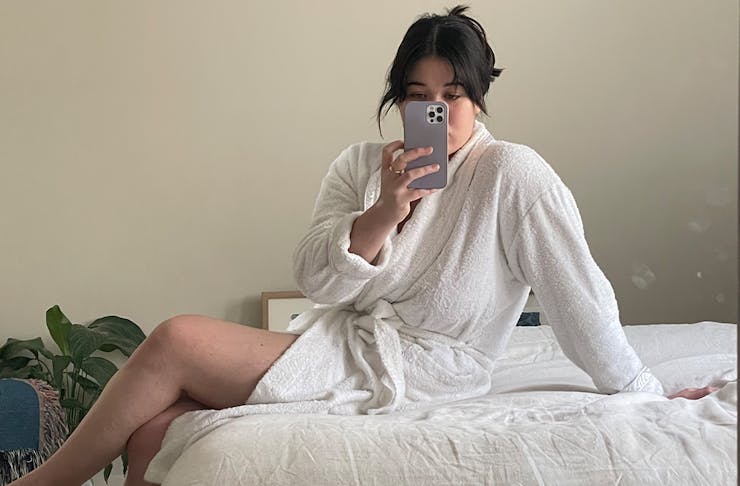 a woman sits on the best in a bathrobe, taking a selfie.
