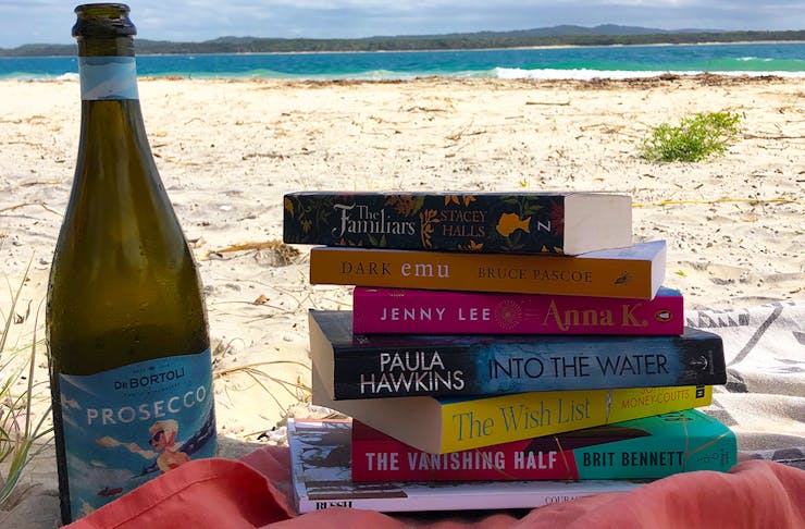 a stack of books next to a bottle of wine on the beach