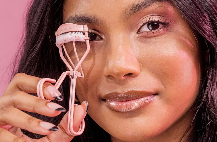 a woman holds up a pink eyelash curler up to her eye.
