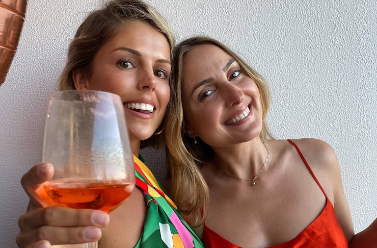 two friend smile at the camera, while one lifts a wine glass in the air.