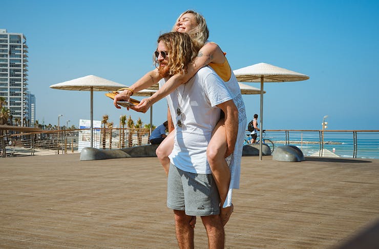 a young man gives a young woman a piggy back. Behind them is a brilliant blue sky, umbrellas and a beach.