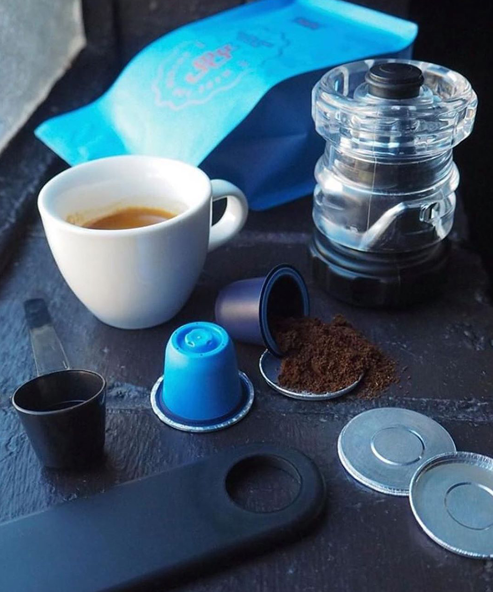 coffee pods and beans sit on a blue benchtop with a cup of coffee in front.