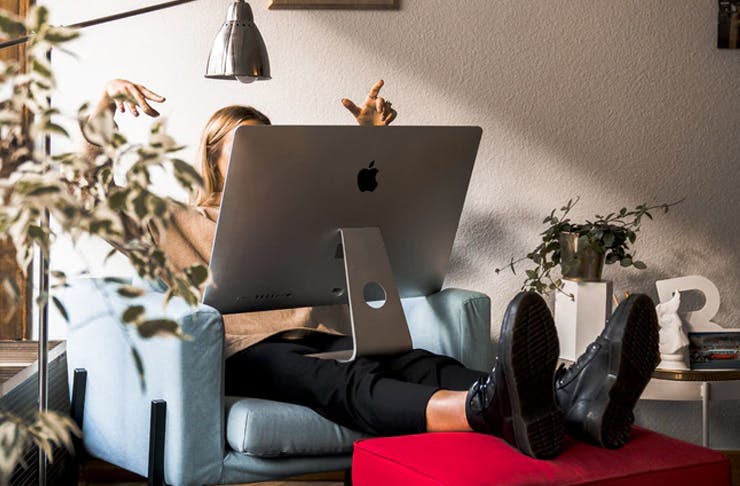 girl sitting in corner of window-lit room on a couch with apple desktop on her legs which are resting on futon