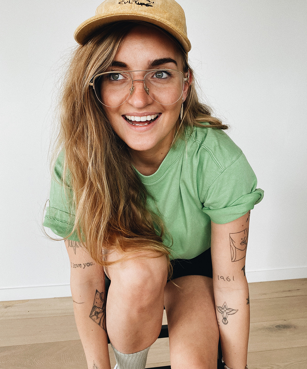 G Flip wearing a green tee, cap and glasses looks off camera with a cheeky smile on her face.