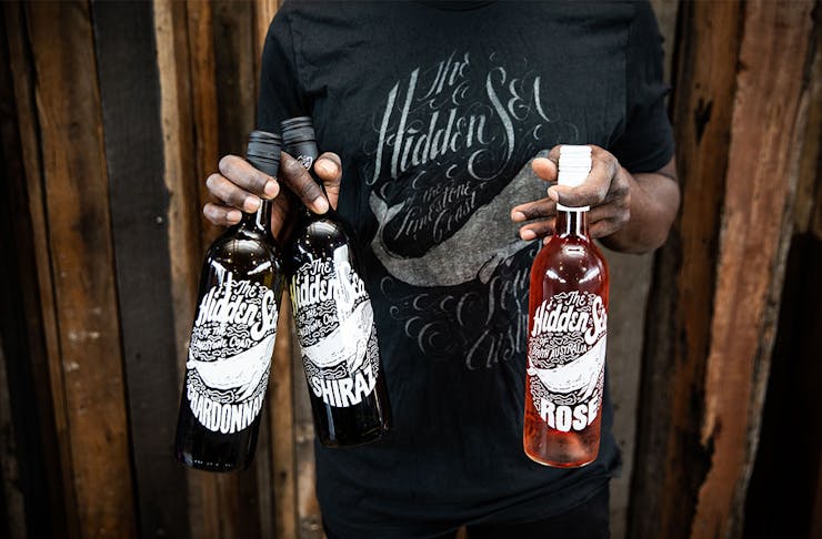 a man holds three bottles of The Hidden Sea wine in his hands. He also wears a t-shirt with The Hidden Sea written on it.