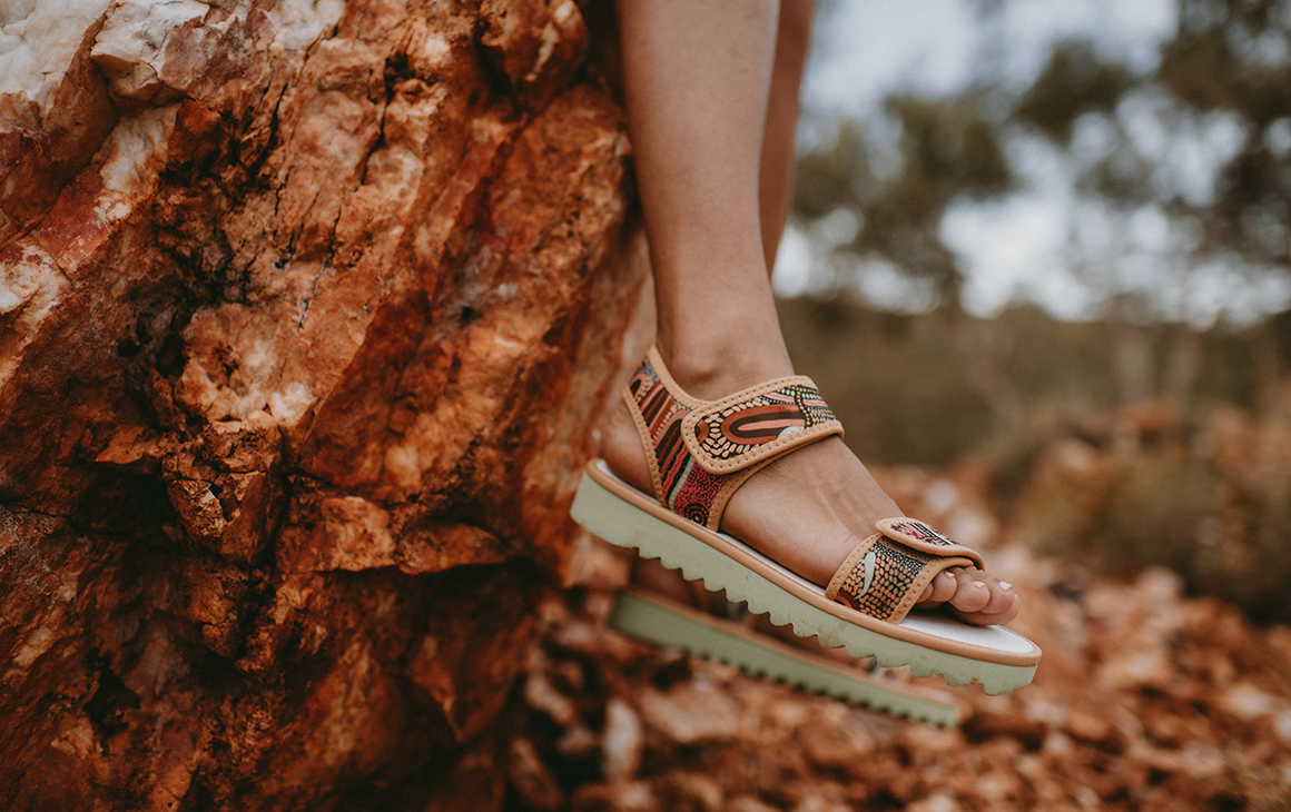 A close up of a pair of TWOOBS X Cungelela Art sandals, against a red rock.